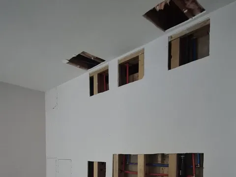 Before: Living room wall and ceiling cut open with dozens of square holes exposing pipes